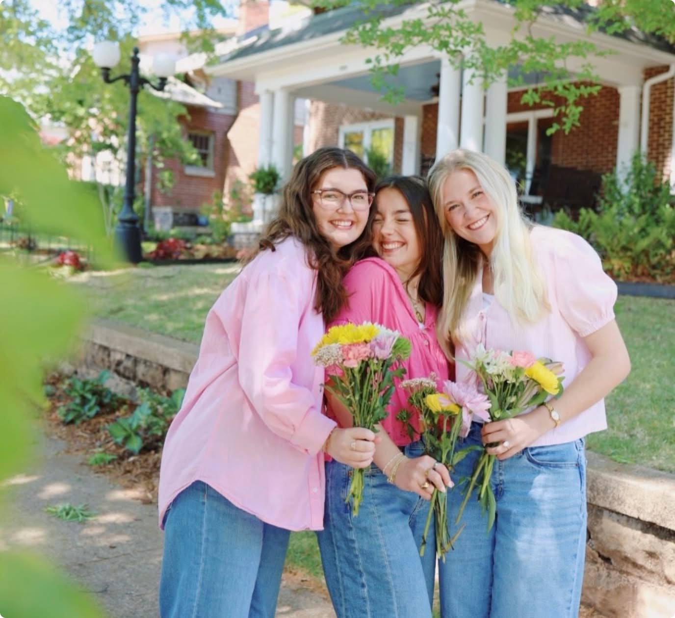 3 sisters smiling holding flowers looking at the camera infront of PMU house